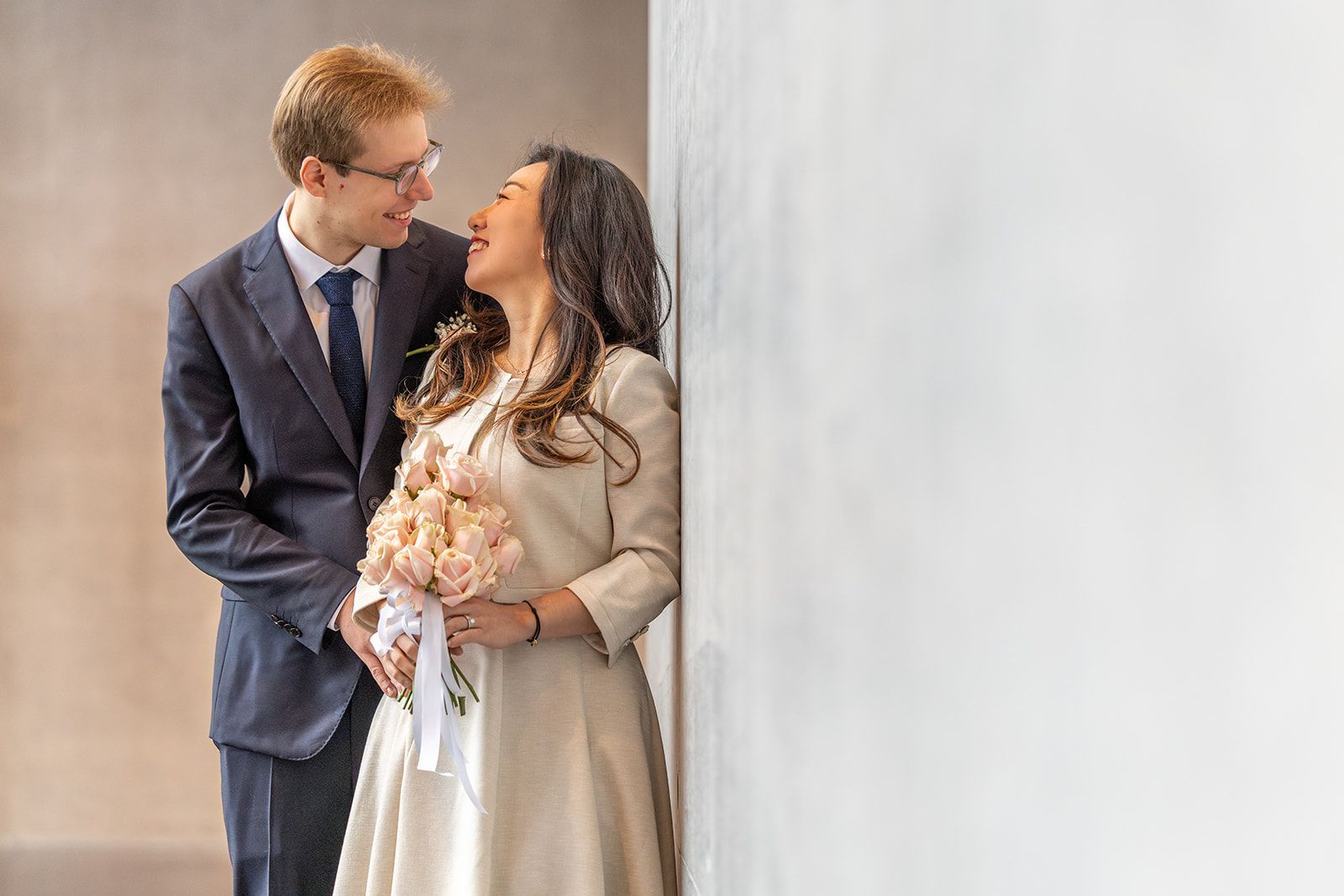 Een lachend stel omhelst elkaar. Een vrouw in een crèmekleurige jurk houdt bloemen vast en leunt tegen een witte muur. Een man in pak glimlacht en staat ernaast.