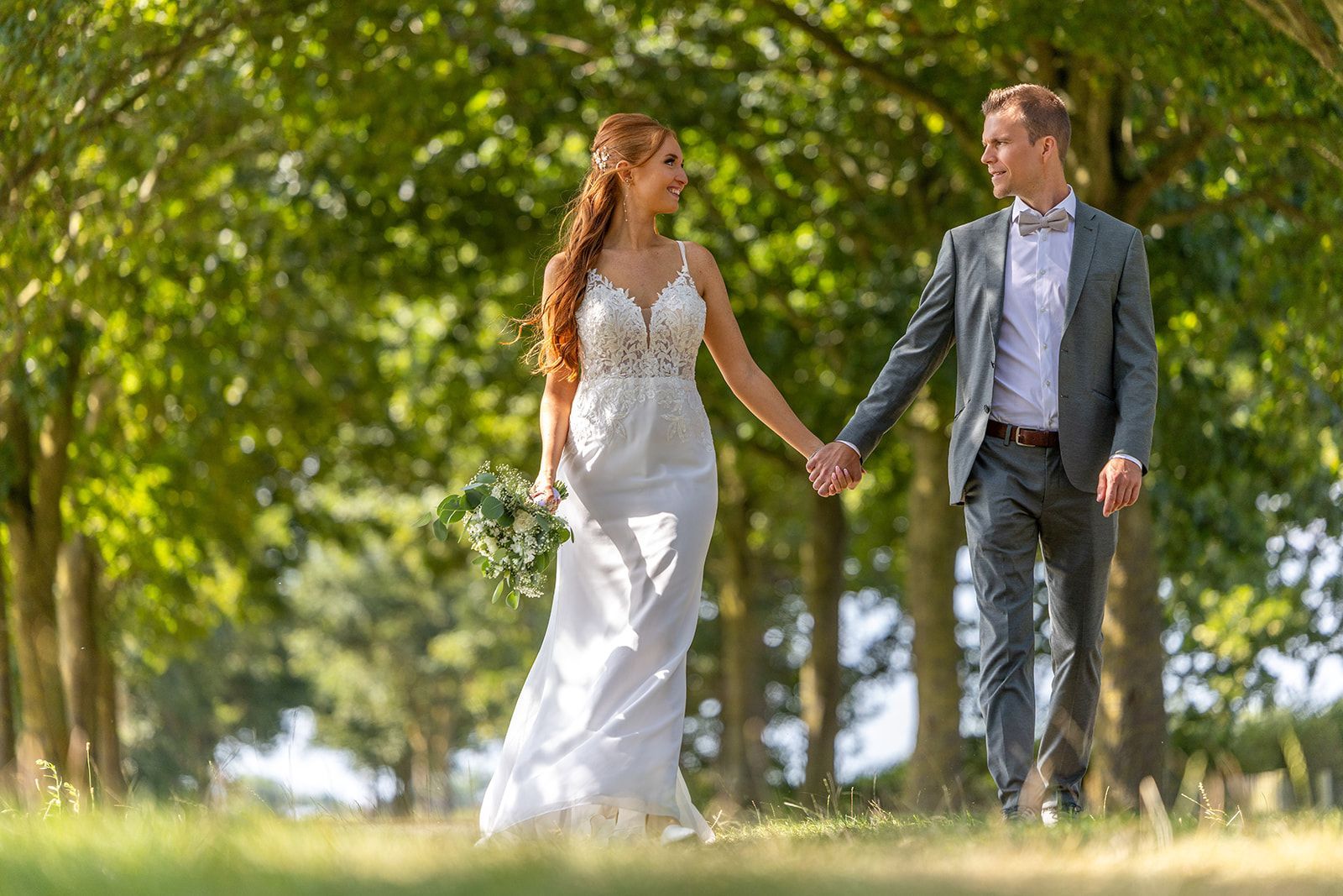 Bruid en bruidegom hand in hand, wandelend in een park onder de bomen. Zij draagt een witte jurk, hij een pak.