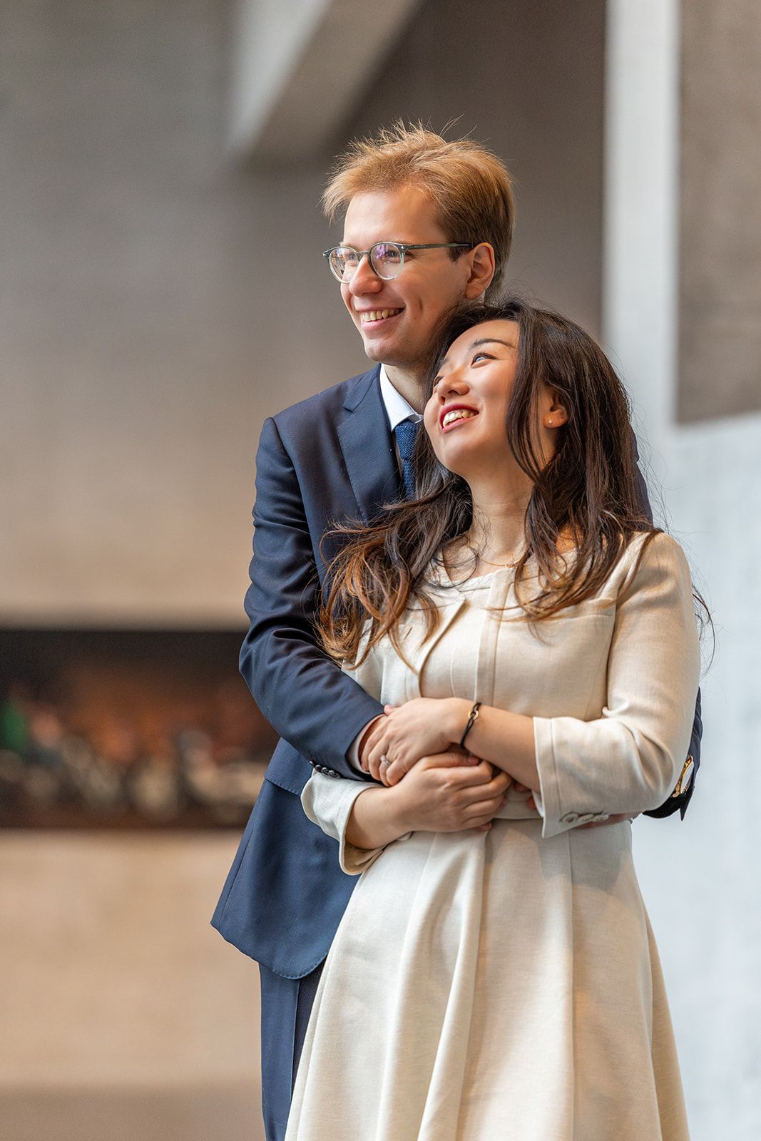Een paar omhelst elkaar, glimlachend. Man in pak, vrouw in crèmekleurige jurk, omhoogkijkend, staand in een gebouw.