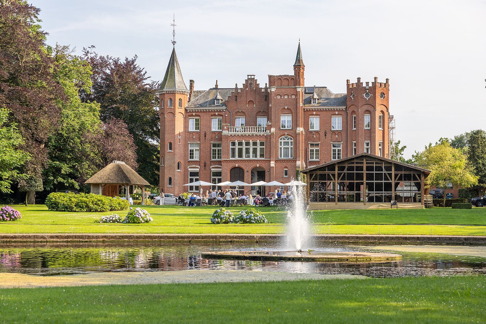 Bakstenen kasteel met fontein op de voorgrond; eettafels op het gazon.