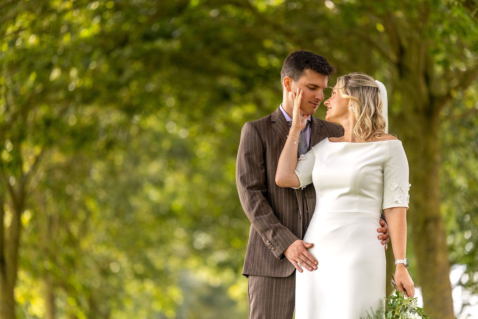 Bride and groom standing in a green park, touching foreheads and holding hands