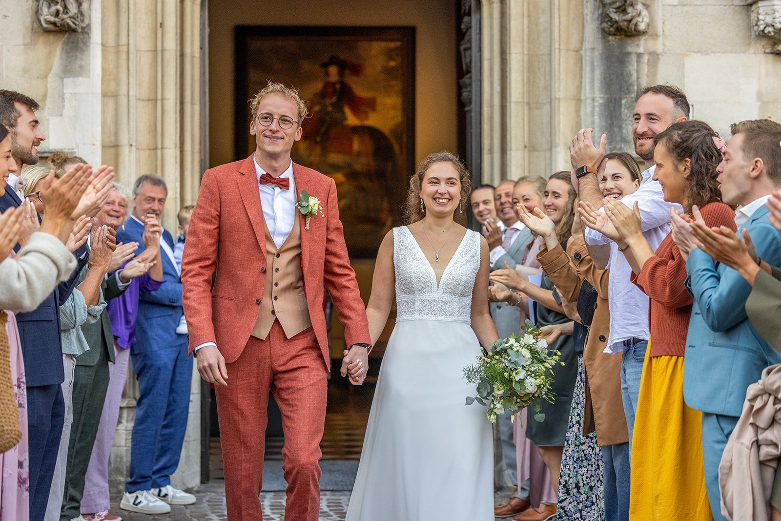 Een pasgetrouwd stel verlaat glimlachend een gebouw, omringd door applaudisserende gasten. De bruidegom draagt ​​een roestbruin pak, de bruid een witte jurk.