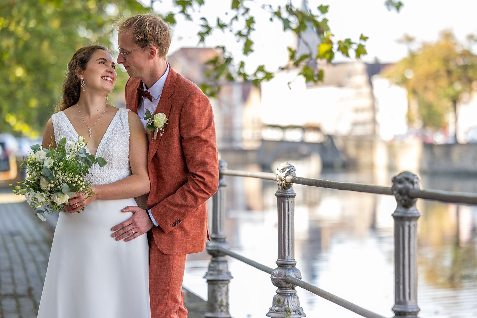 Bruid en bruidegom staan ​​bij een waterpartij. Zij draagt ​​een witte jurk, houdt bloemen vast en kijkt hem aan. Hij draagt ​​een roestkleurig pak en glimlacht.