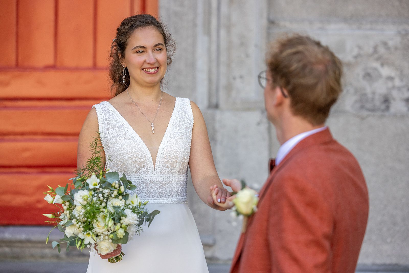 Bruid in witte jurk lacht, houdt boeket vast en kijkt naar bruidegom in rood pak bij oranje deur.