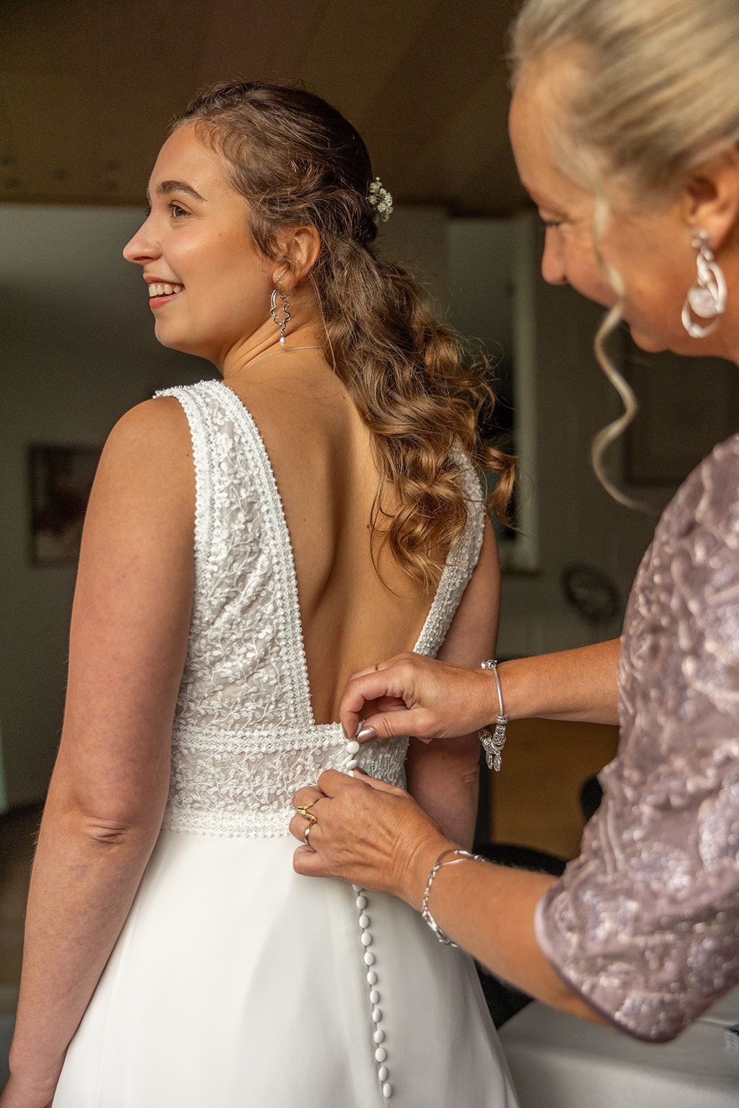 Bruid lacht terwijl vrouw de achterkant van haar trouwjurk vastmaakt.