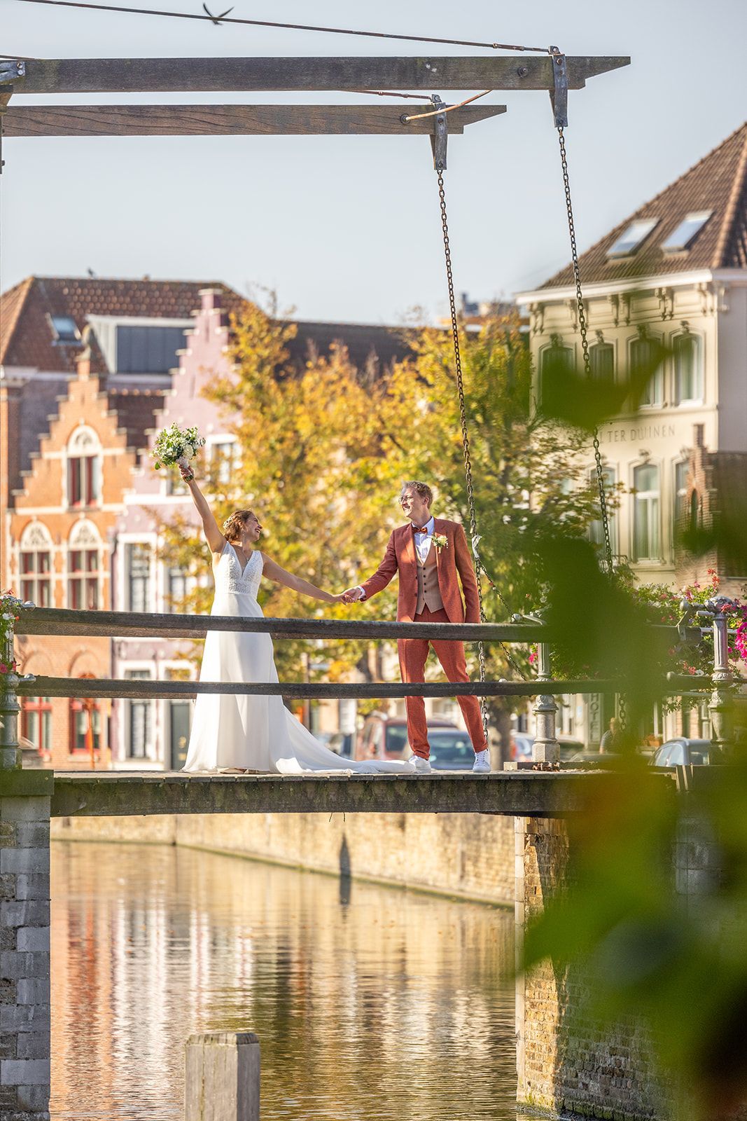Bruid en bruidegom op een brug over het water, hand in hand, poserend voor een foto. Historische gebouwen op de achtergrond.