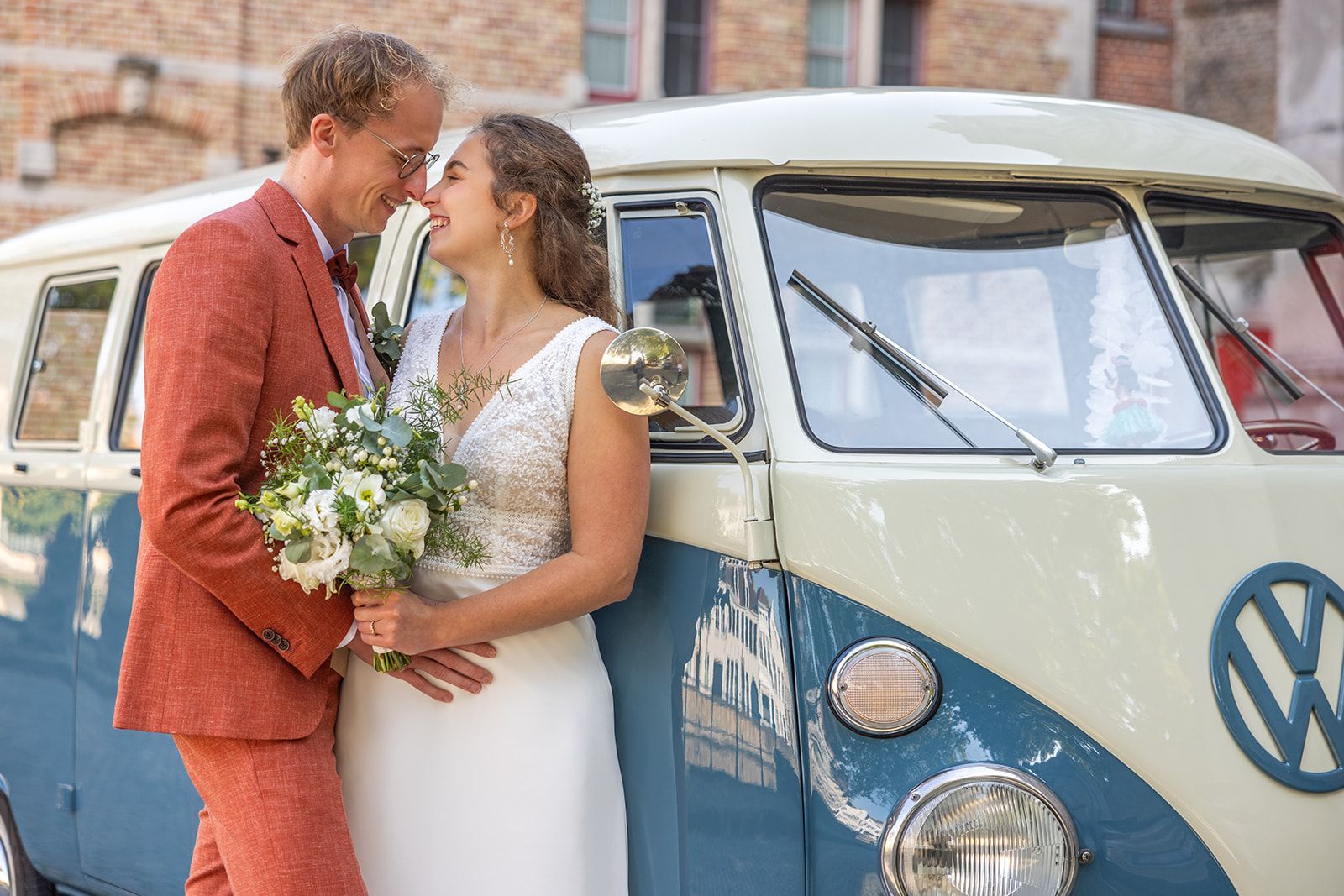 Een pasgetrouwd stel omhelst elkaar naast een vintage blauw-wit Volkswagenbusje en houdt bloemen vast.