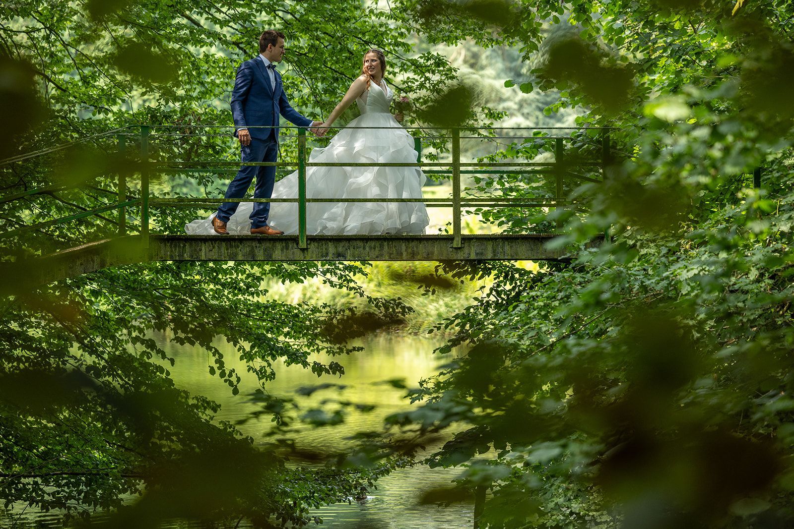Een bruidspaar op een groene brug over een kreek, omgeven door weelderige groene bomen.