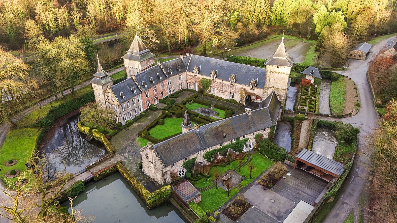 Luchtfoto van een kasteel met een slotgracht, tuinen en omringende bomen. Grijs stenen gebouw, groen gras en bruine bomen.
