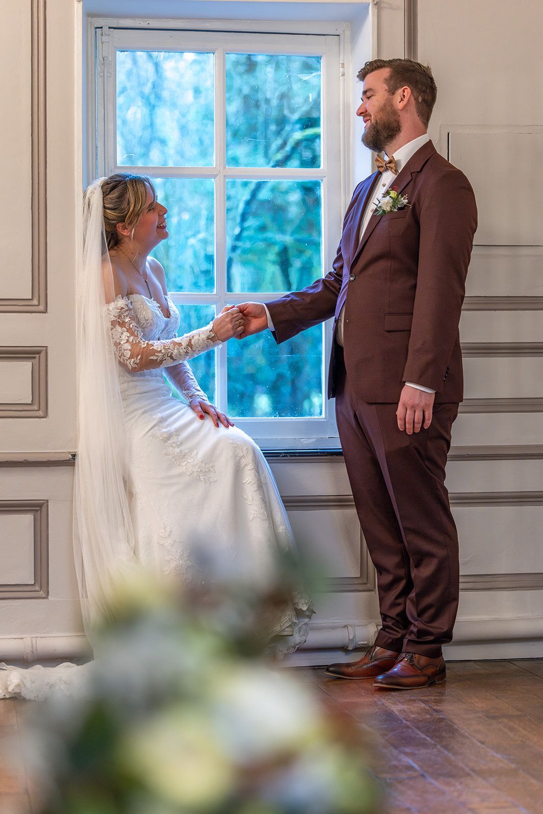 De bruid in de witte jurk zit bij het raam, hand in hand met de bruidegom in het bordeauxrode pak, en lacht.