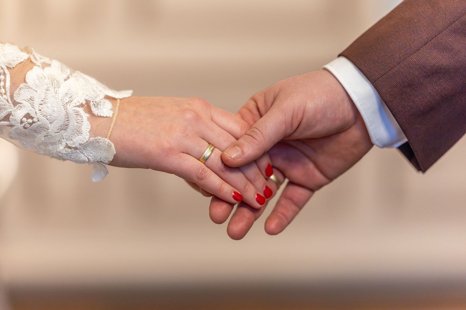Een stel houdt elkaars hand vast en draagt ​​trouwringen. De hand van de vrouw heeft een kanten mouw, de hand van de man draagt ​​een bruin pak en rode nagellak.
