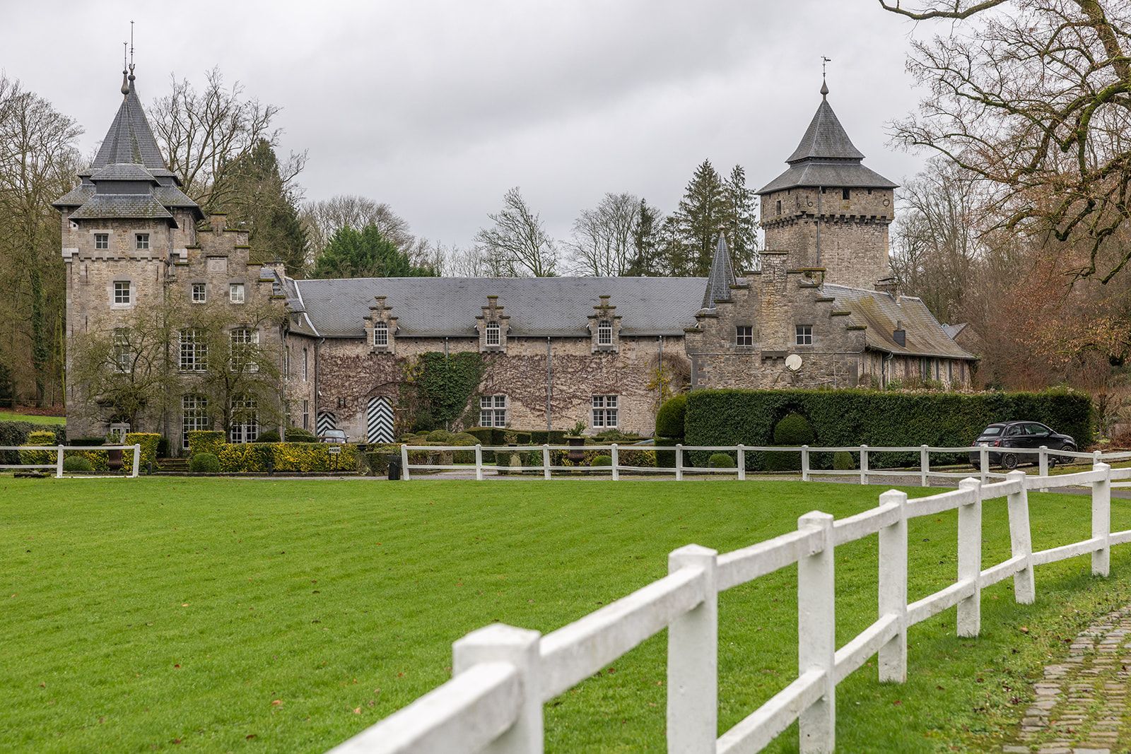 Stenen kasteel met twee torens, een lang dak en een wit hek voor een groen veld.