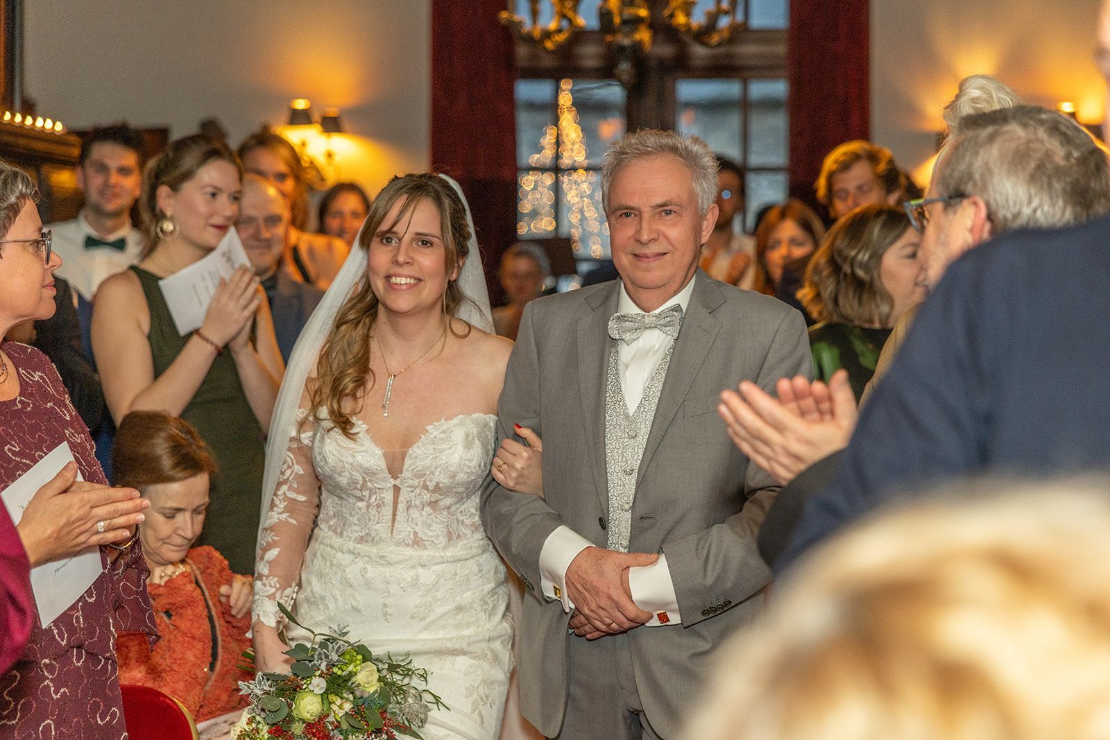 Bruid en man in grijs pak lopen glimlachend, arm in arm, door de menigte en applaudisseren. Binnen met rode muur.