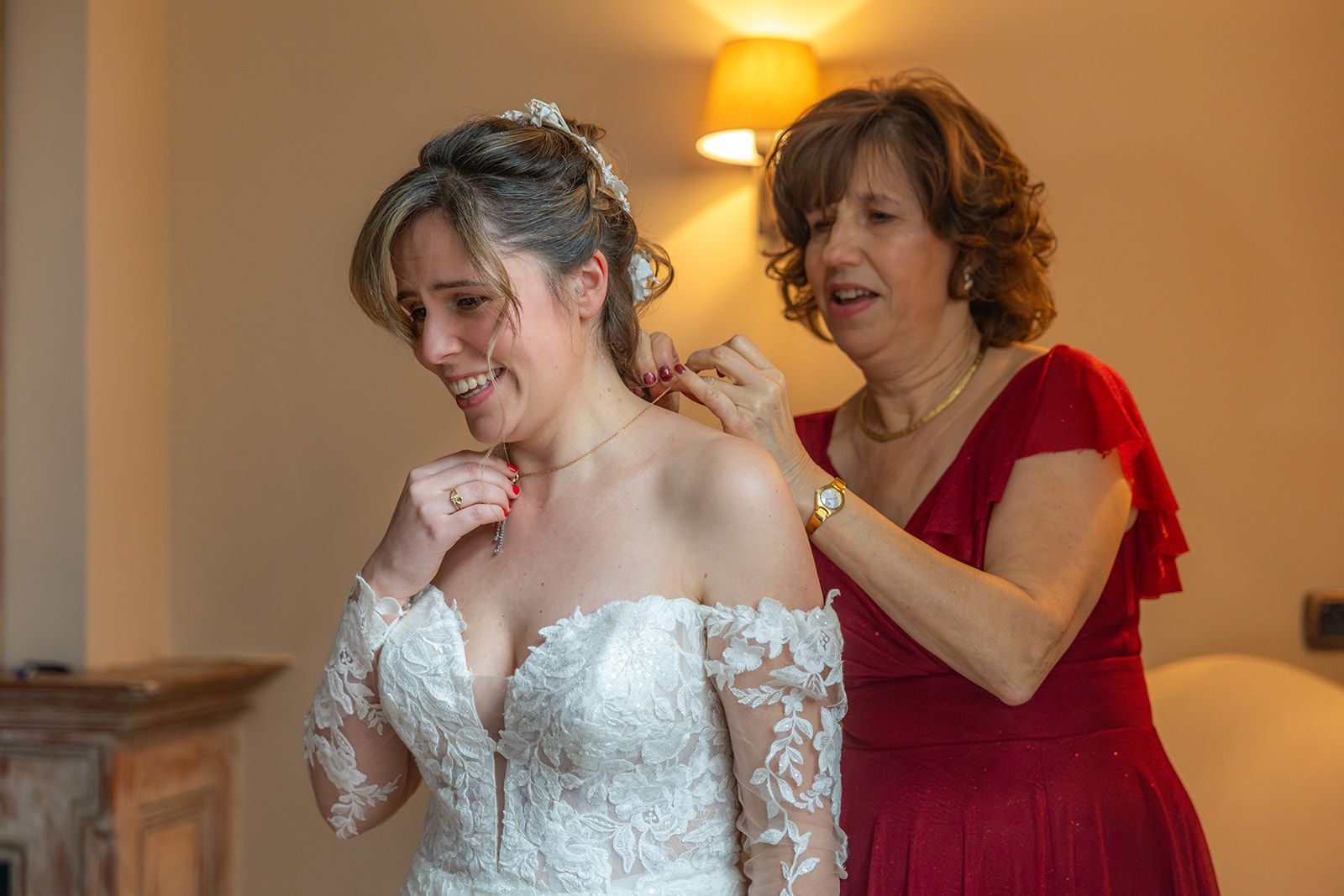 Bruid in trouwjurk krijgt haar ketting rechtgezet door een vrouw in een rode jurk. Interieur.