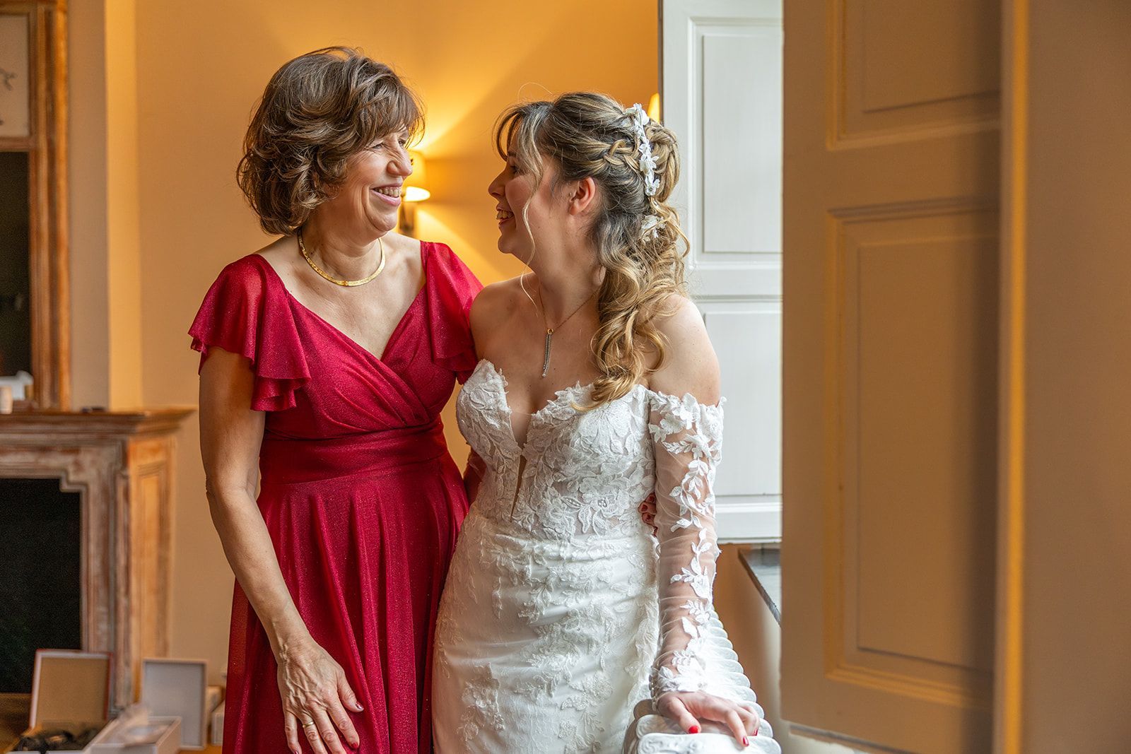 Bruid in witte jurk lacht naar vrouw in rode jurk. Interieurfoto vlakbij raam.