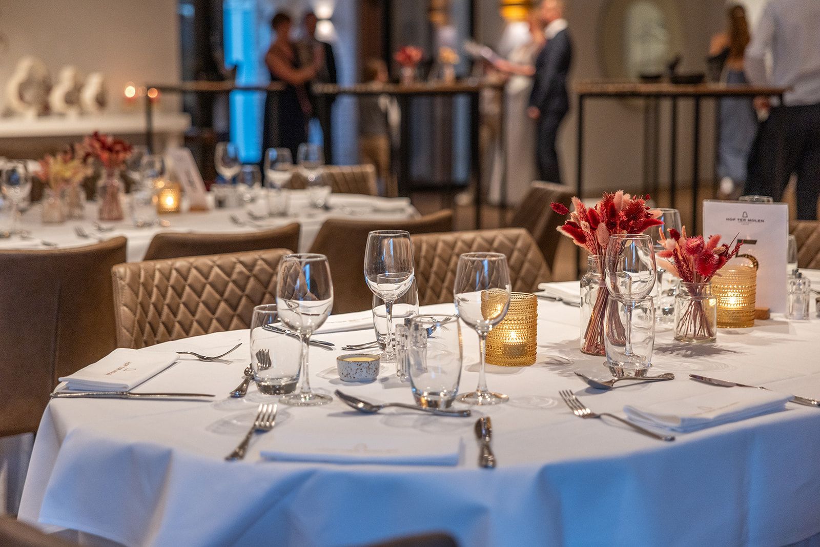 Formeel gedekte eettafel voor een speciale gelegenheid, met glaswerk, bloemen en een tafelnummer. De andere tafels en mensen zijn vaag op de achtergrond te zien.