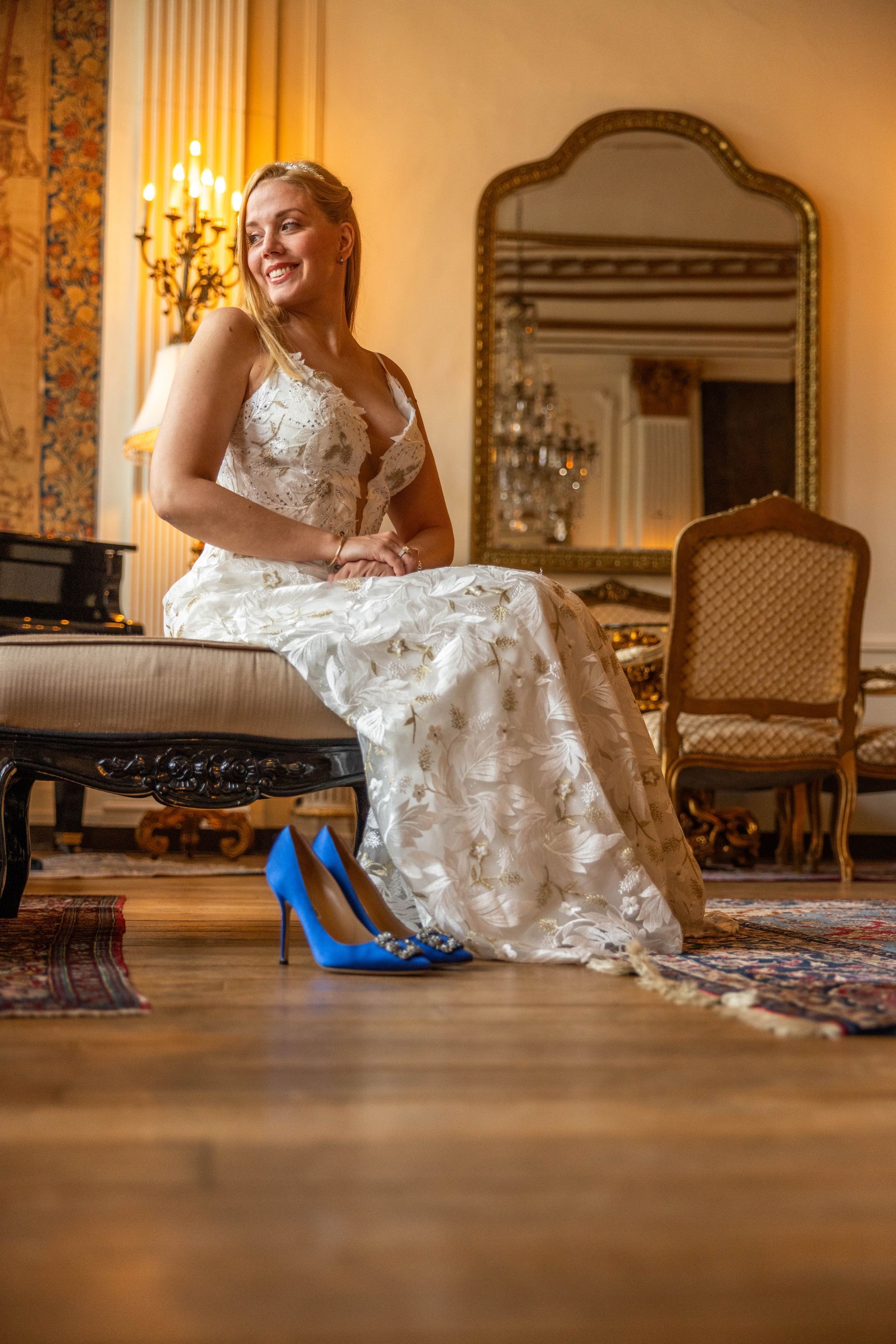 Een bruid in een trouwjurk zit glimlachend, met blauwe hakken op de voorgrond. Een elegante kamer met spiegel, piano en tapijten.