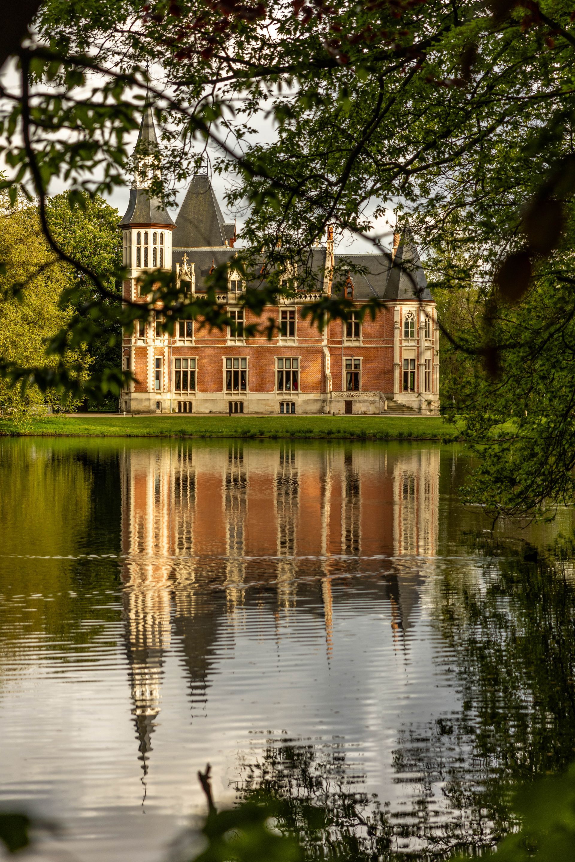 Bakstenen landhuis weerspiegeld in stilstaand water, omgeven door bomen.