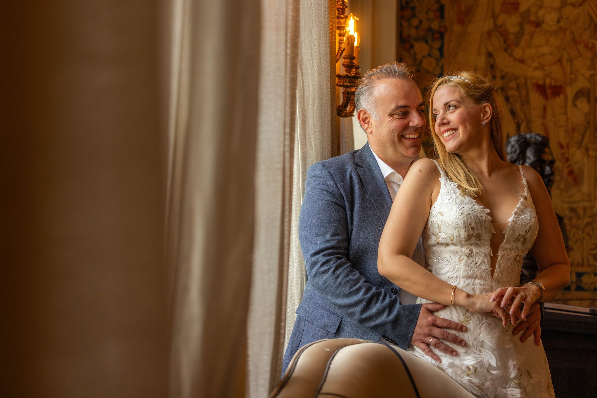 Man and woman embrace in front of a window, smiling. He wears a blue blazer, she wears a white dress.