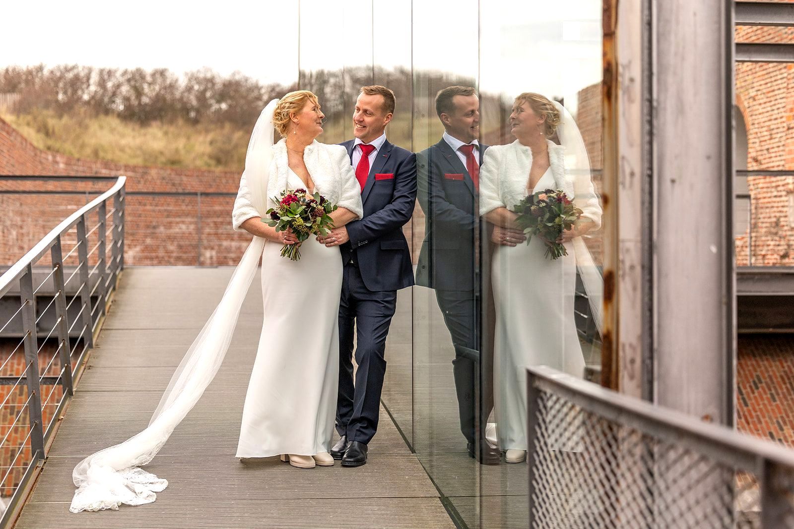 Bruid en bruidegom op een brug, glimlachend en elkaar aankijkend. Bruid draagt een sluier, bruidegom een rode stropdas. Weerspiegeling op glas.