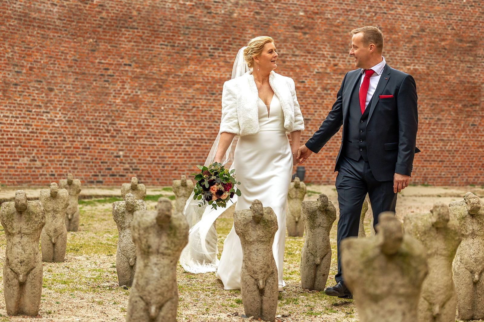 Bruid en bruidegom lopen hand in hand langs stenen sculpturen. Bruid in witte jurk, bruidegom in pak, bakstenen muur op de achtergrond.