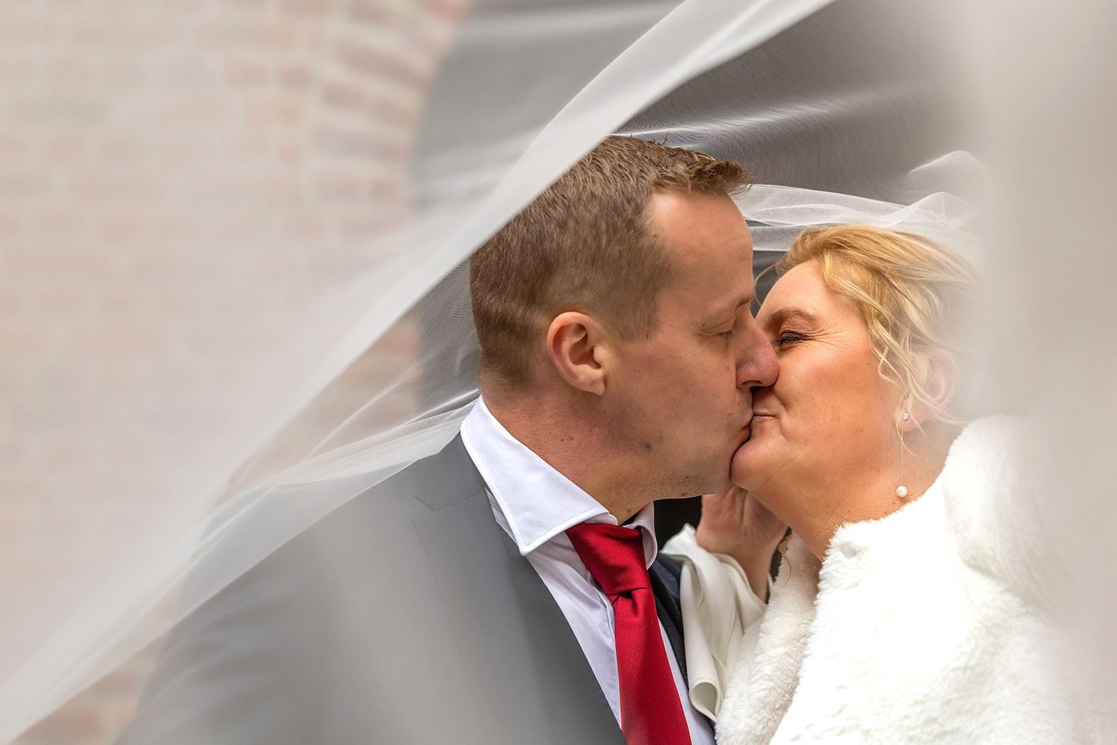 Kussend stel onder een sluier; man in pak met rode stropdas, vrouw in witte jurk, bakstenen achtergrond.