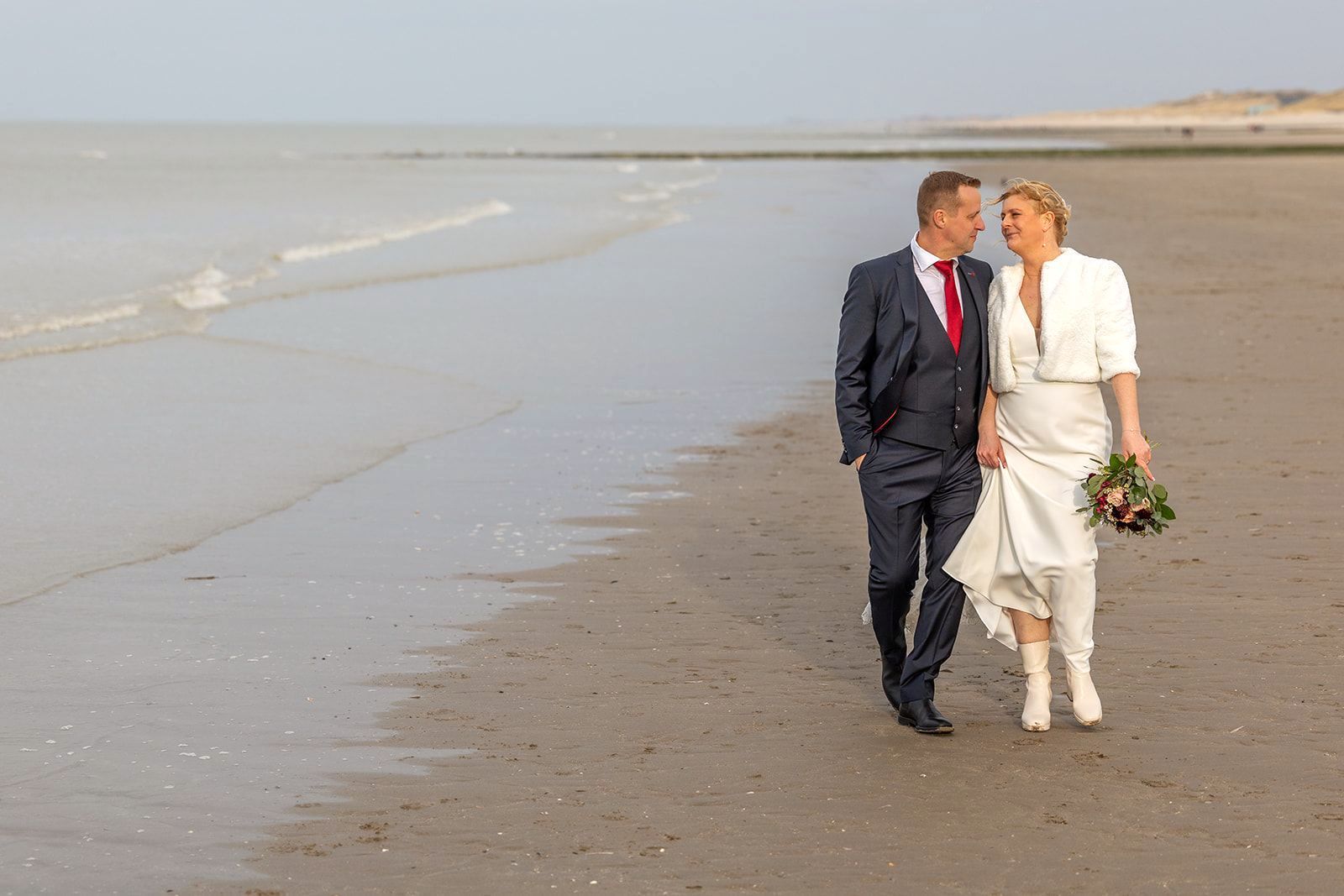 Een stel loopt op het strand, man in pak, vrouw in trouwjurk, hand in hand, lachend.