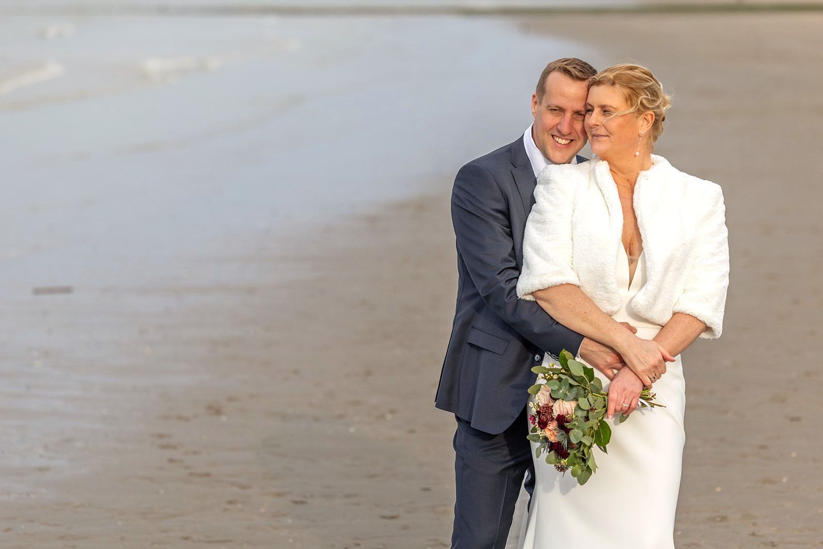 Een stel in bruidsjurk omhelst elkaar op het strand; de man in pak, de vrouw in een witte jurk, ze houden bloemen vast en lachen.