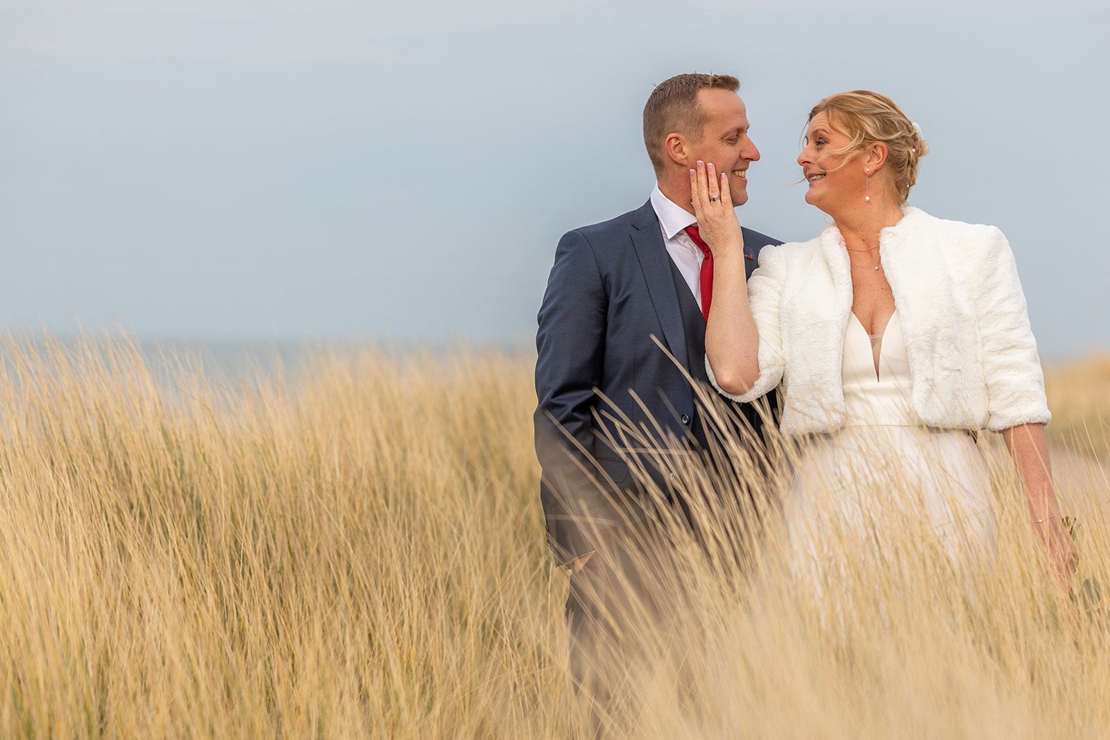 Een bruidspaar omhelst elkaar te midden van hoog gras en kijkt elkaar aan. Man in pak, vrouw in witte jurk, glimlachend.