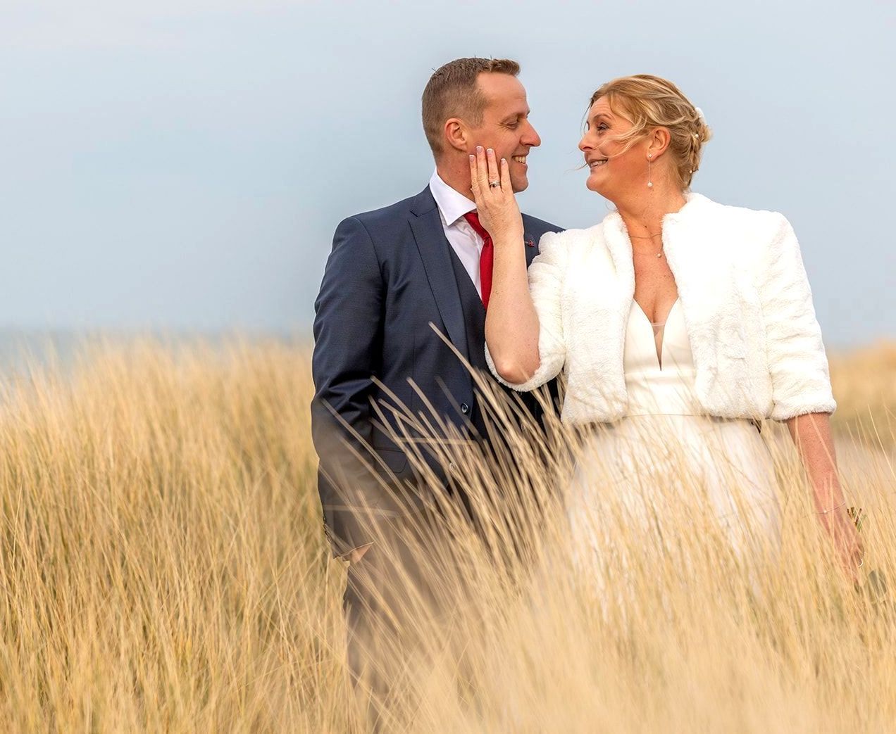 Bruid en bruidegom in formele kleding, lachend naar elkaar in een hoog grasveld, met een blauwe lucht op de achtergrond.