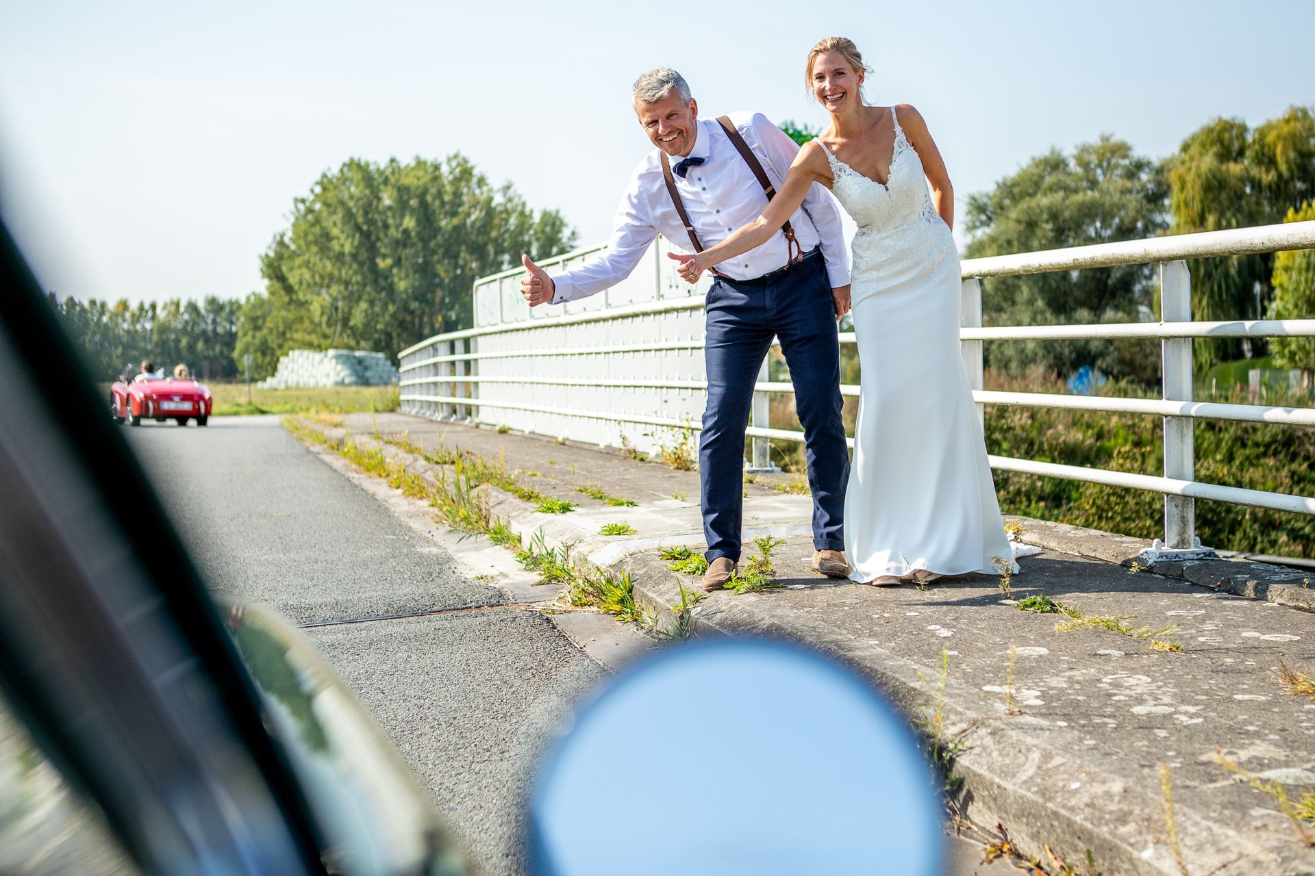 Bruid en bruidegom liften op een brug en zwaaien naar een rode auto. Zonnige dag.
