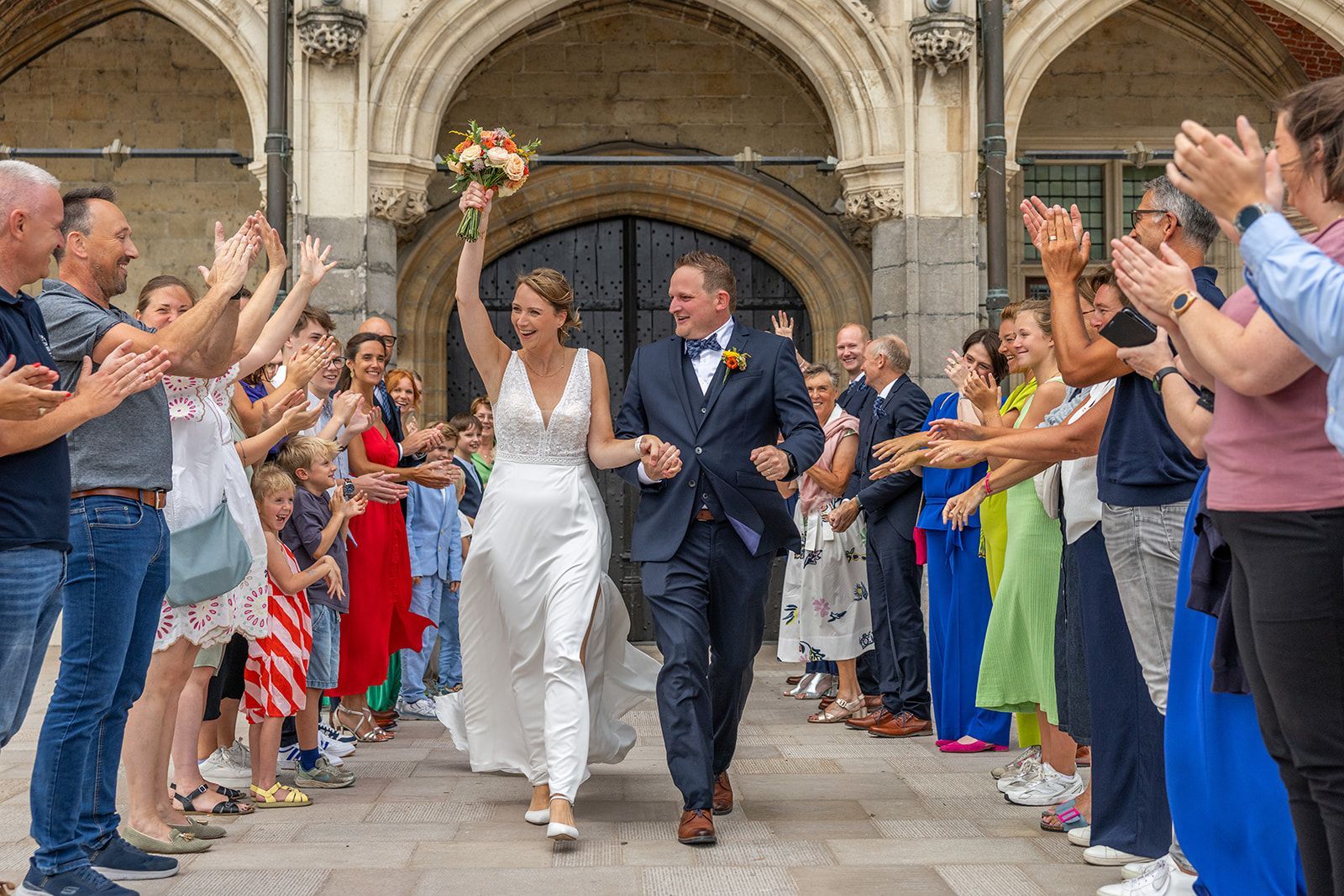 Newlyweds cheered by wedding guests as they exit a building. Bride raises bouquet, groom smiles, everyone applauds.