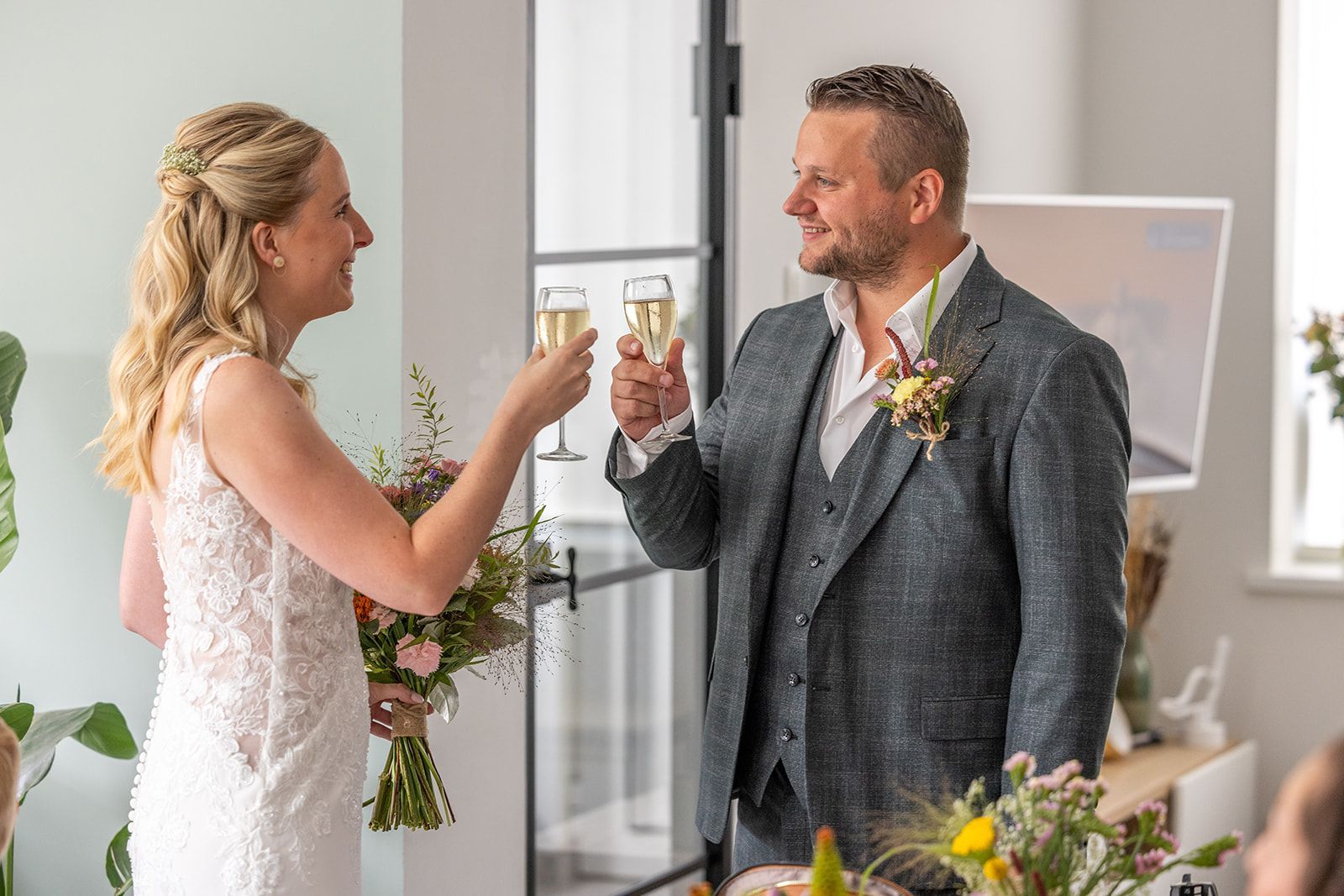 Bruid en bruidegom toosten met champagne; binnen, met bloemstukken.