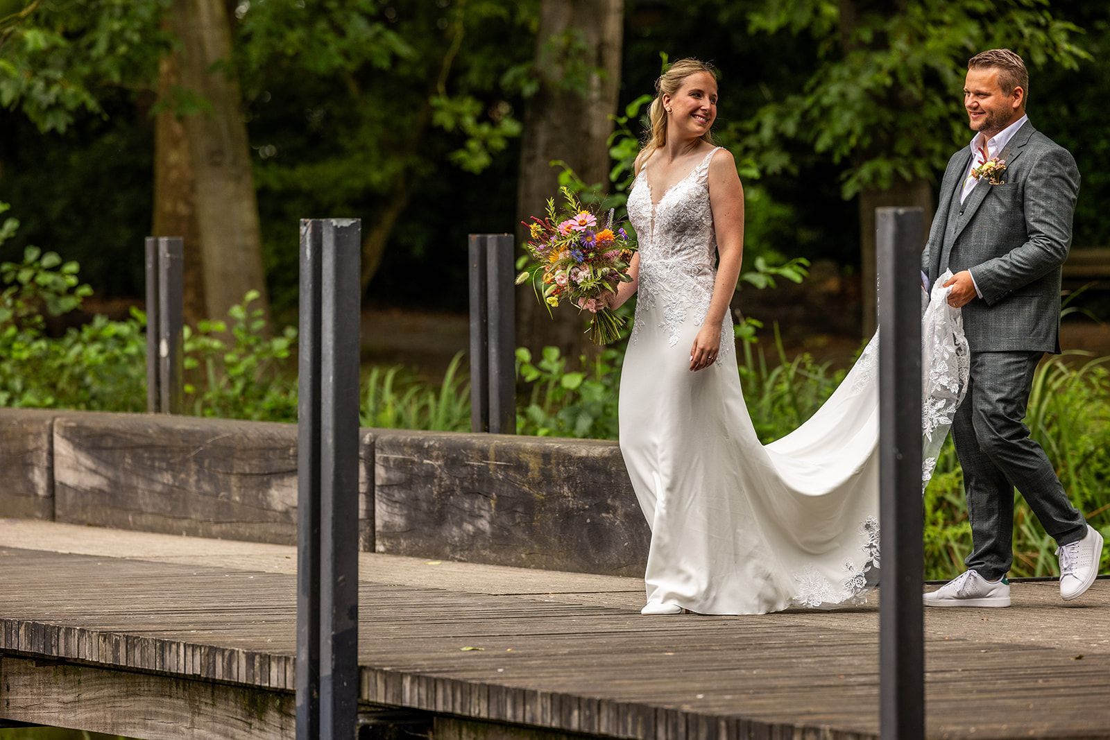 Bruid in witte jurk met sleep, glimlachend naar bruidegom in grijs pak, lopend op houten pier, met boeket in de hand.