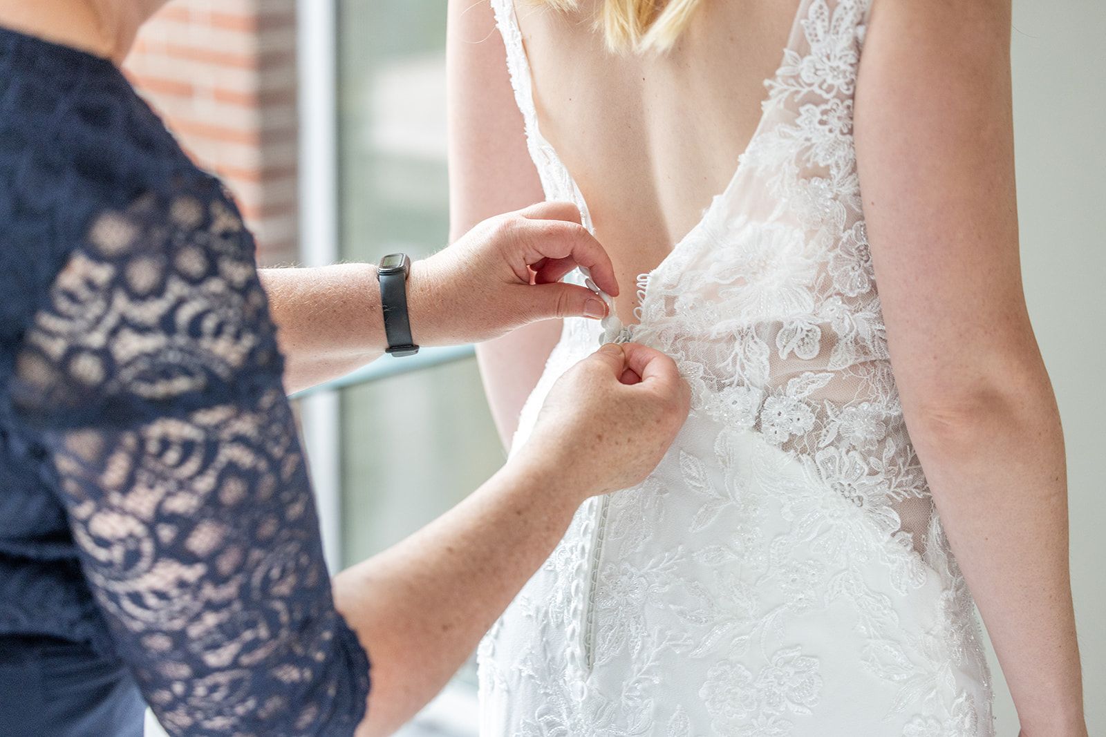Een vrouw in een trouwjurk, met haar rug naar de kijker, laat haar jurk door iemand anders dichtknopen.