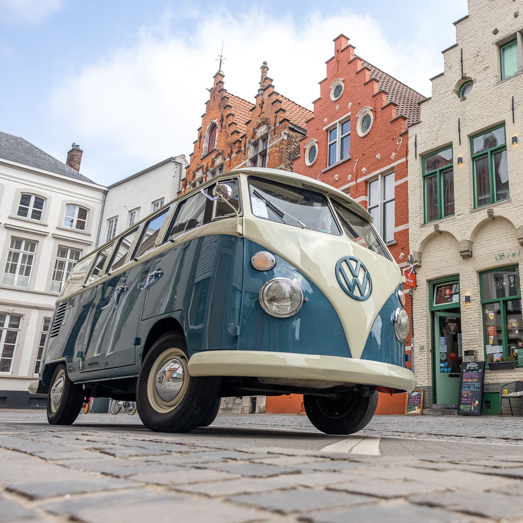 Een blauw-witte klassieke Volkswagenbus geparkeerd op een geplaveide straat, met historische gebouwen op de achtergrond.