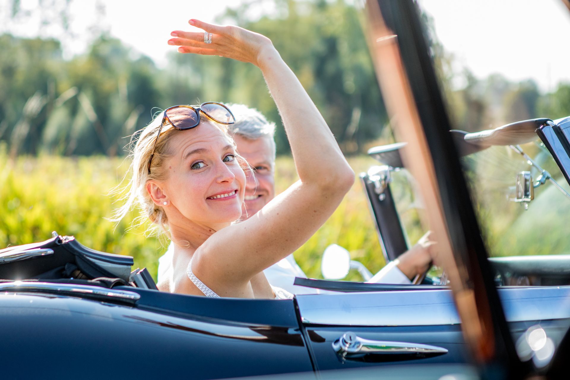 Een vrouw zwaait vanuit een klassieke cabriolet en lacht naar de camera, met een man naast haar. Zonnig weer.