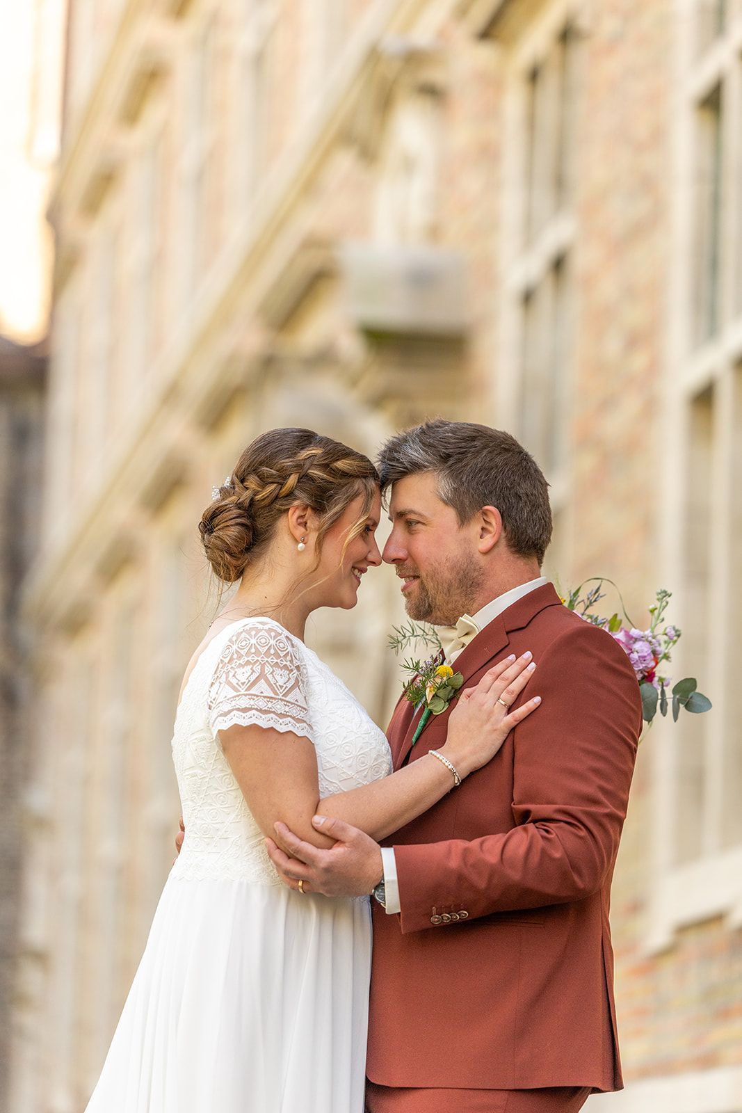 Een glimlachend bruidspaar, voorhoofd aan voorhoofd, bij een stenen gebouw. De bruidegom draagt een roestbruin pak, de bruid een witte jurk en houdt bloemen vast.