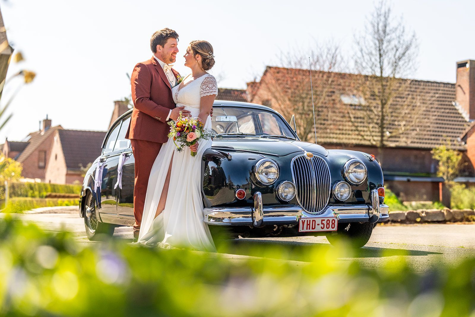 Bruidspaar poseert bij een zwarte vintage auto, met bloemen in de hand, op een zonnige locatie.
