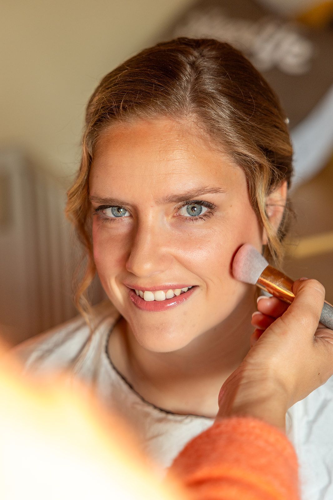 Visagiste brengt blush aan op de wang van een vrouw. De vrouw glimlacht, lichte huid, blond haar, binnen.