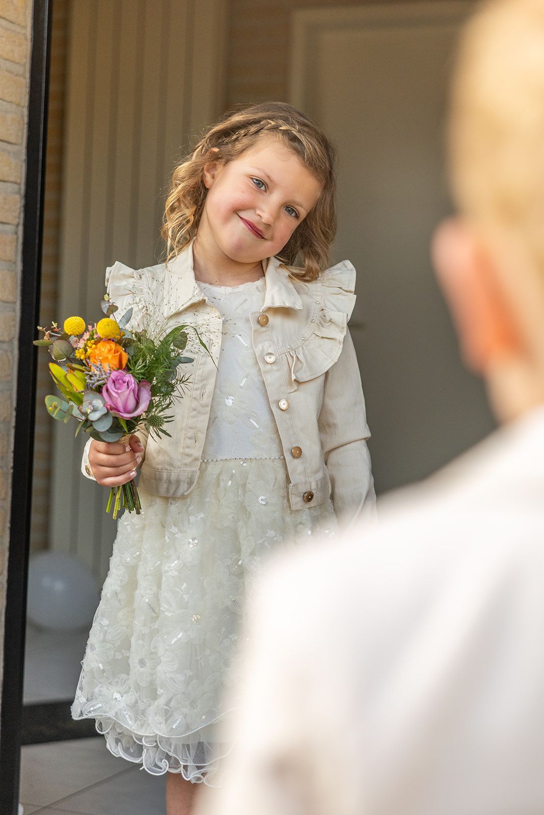 Meisje in een witte jurk en jas houdt bloemen vast en kijkt naar iemand buiten beeld.