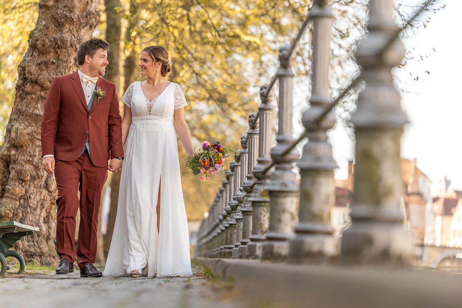 Pasgetrouwden lopen hand in hand buiten. Man in roestbruin pak, vrouw in witte jurk met bloemen, langs een pad.