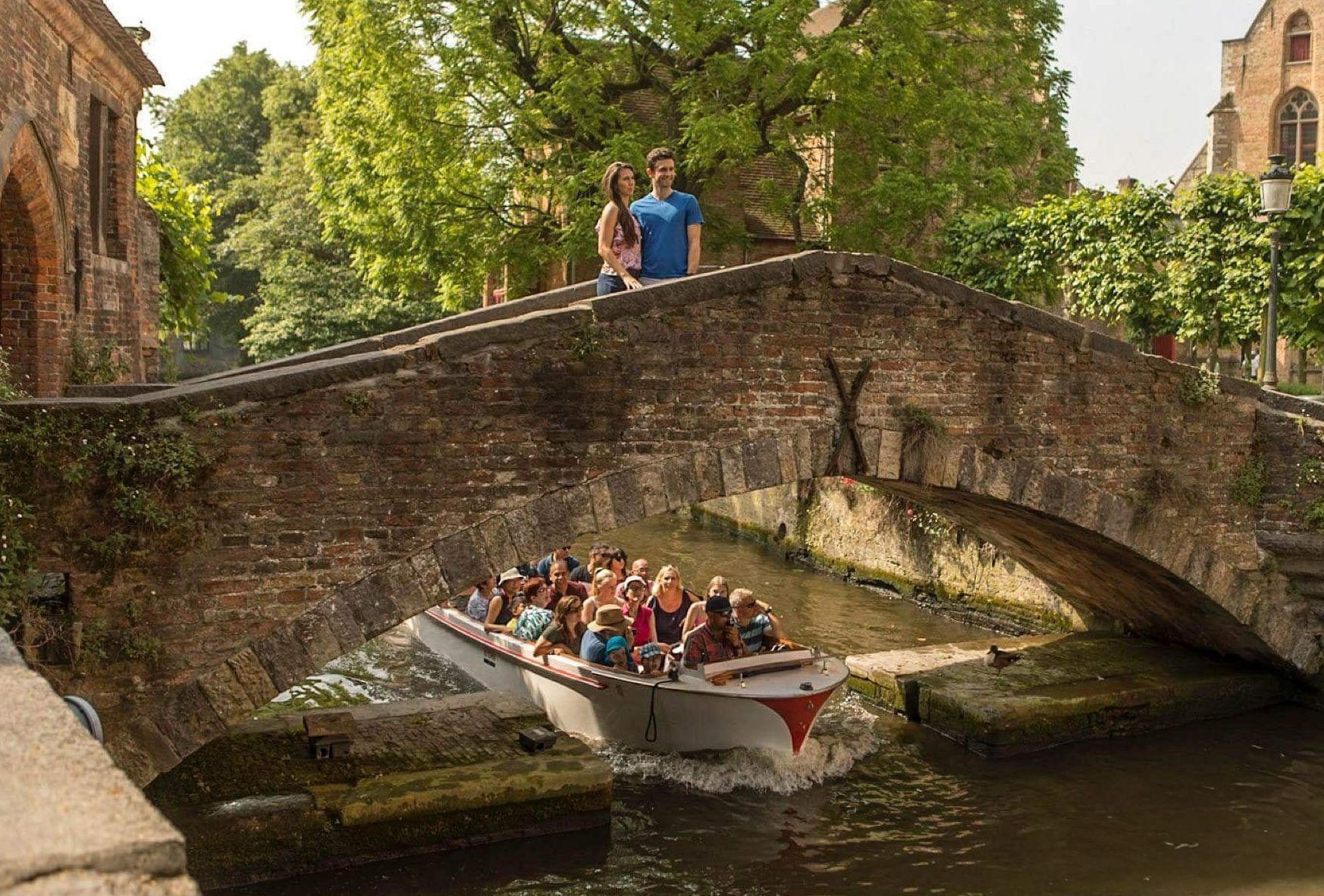 Paar op stenen brug, boot met toeristen die beneden passeren op kanaal, Brugge.