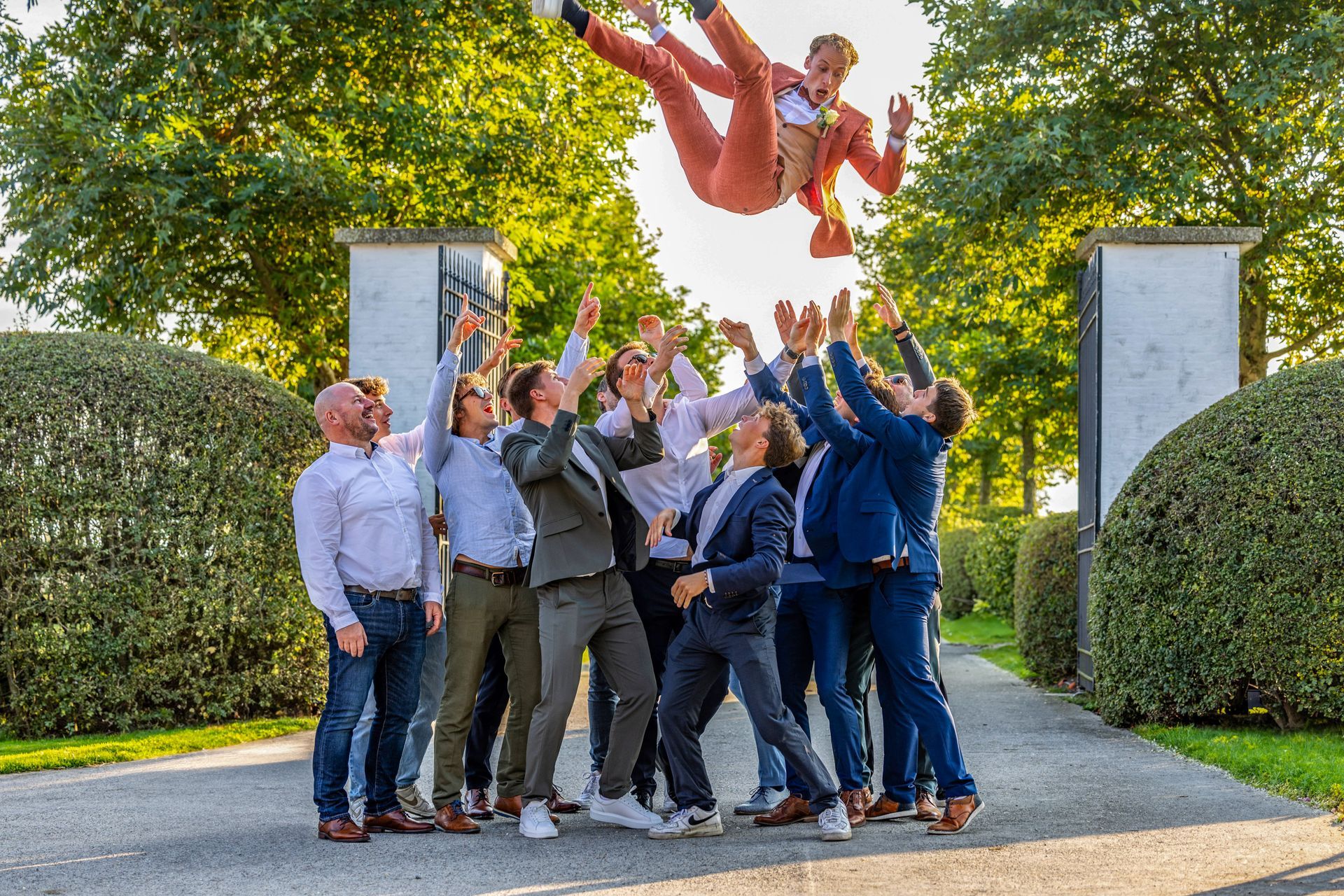 Een groep mannen gooit een man in een zalmkleurig pak in de lucht bij een stenen boog.