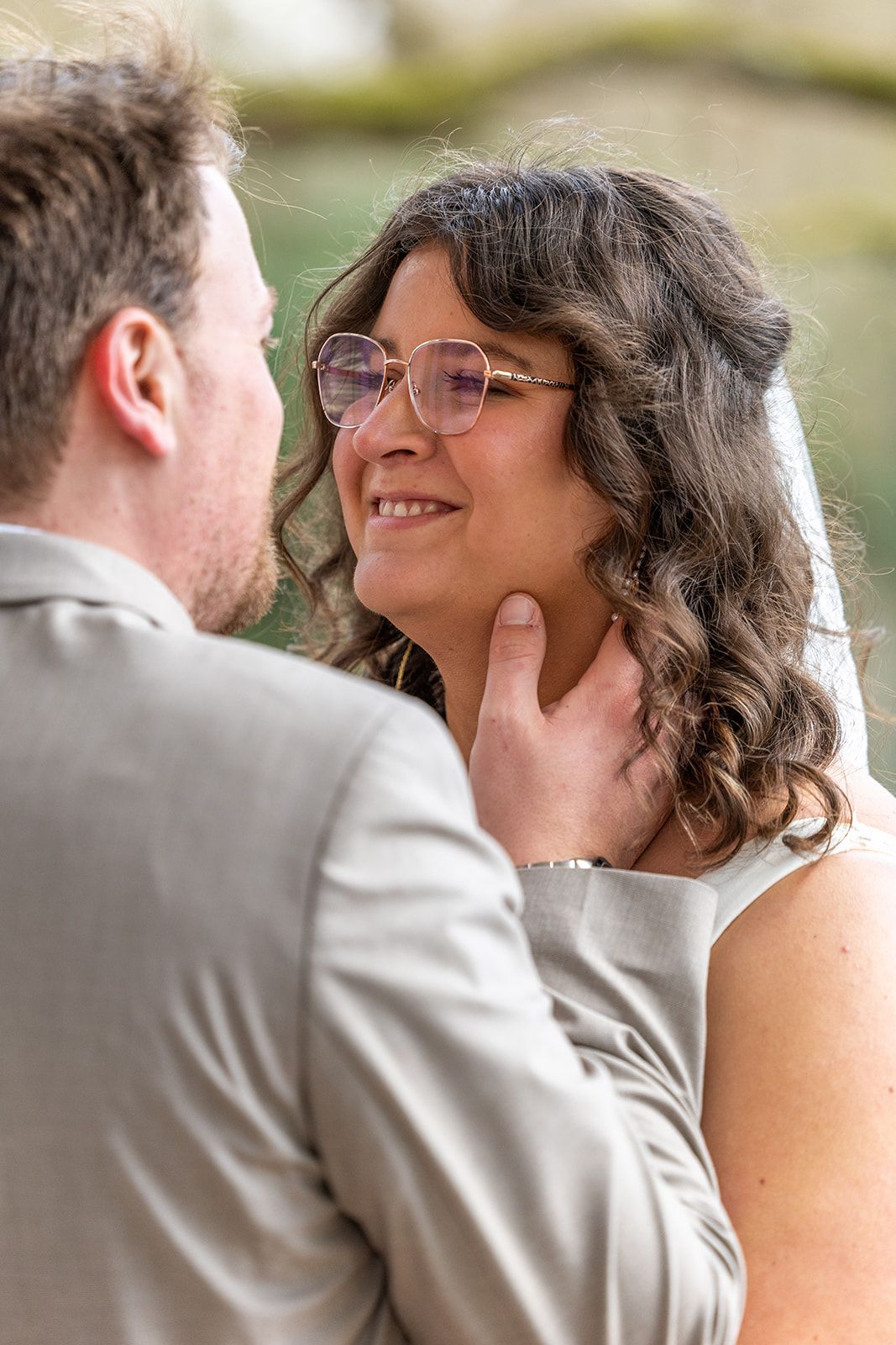 Een stel omhelst elkaar op het strand; de vrouw in de witte jurk houdt een boeket vast, de man in het beige pak raakt haar gezicht aan, met de oceaan op de achtergrond.