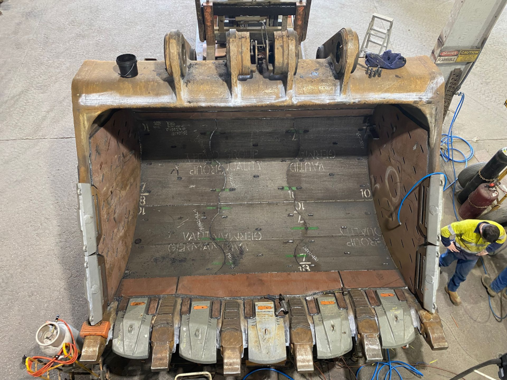 Large excavator bucket, open and facing the viewer, with visible wear and tear. A worker is seen nearby— North Queensland Hard Facing (NQHF) In Fredericksfield, QLD