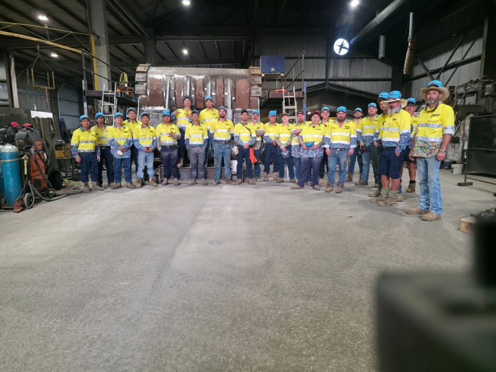 Group of workers in yellow safety vests and blue hats pose in a workshop— North Queensland Hard Facing (NQHF) In Fredericksfield, QLD