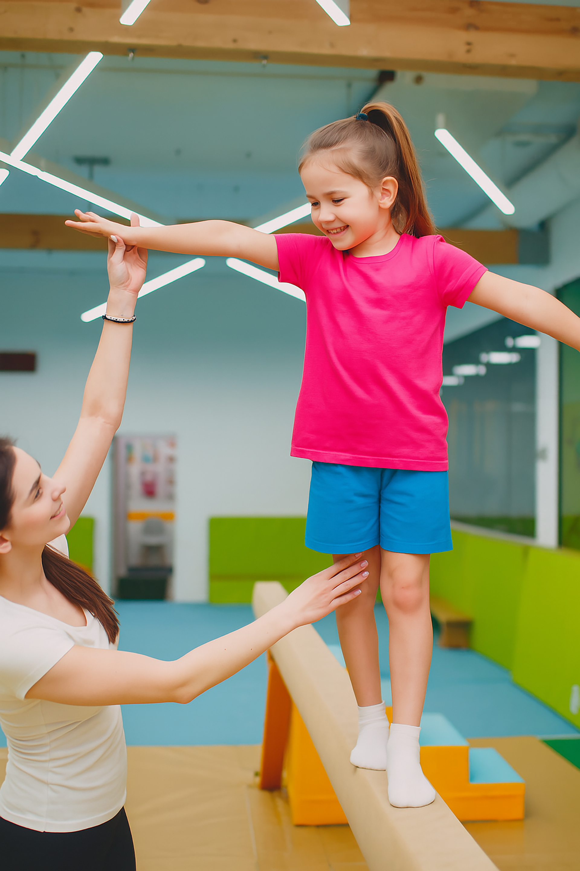 Girl balancing on beam, assisted by woman, in a gymnasium. Girl wears pink shirt and blue shorts.
