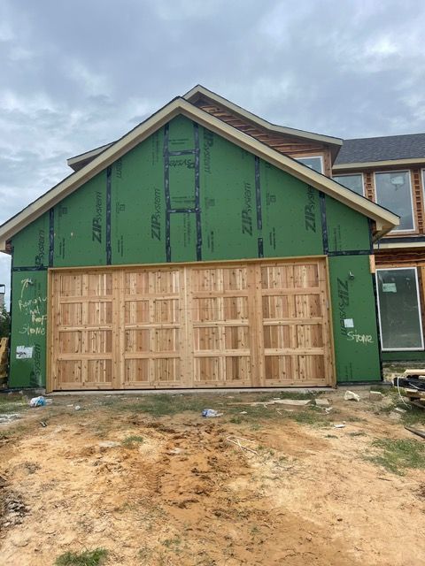Exterior view of a house under construction; wooden garage door. Green sheathing and dirt in the foreground.