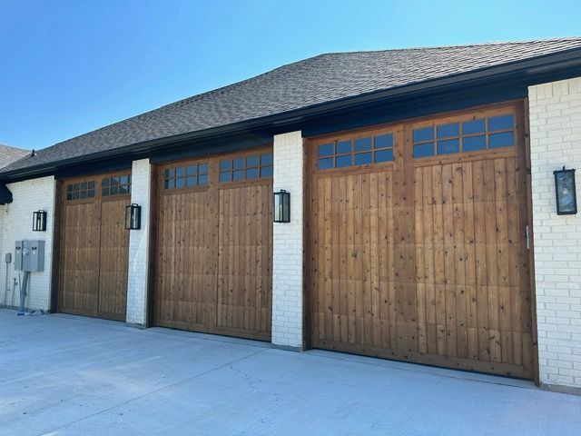 Three wooden garage doors with windows, flanked by white brick and black lights, under a dark roof.